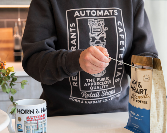 Person scooping cofffee wearing a dark grey sweatshirt that says restaurants, automats, cafeterias, retail shops. Horn & Hardart Est 1888