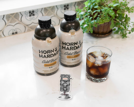 Two bottles of Horn & Hardy Cold Brew coffee with ice, a glass of iced coffee, and a small magnet on a white countertop with a tiled backsplash and a potted plant in the background.