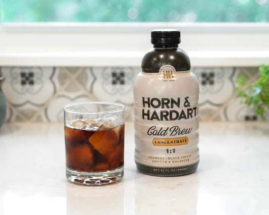 A bottle of Horn & Hardart Cold Brew coffee next to a glass of iced coffee on a white marble countertop in front of a window.