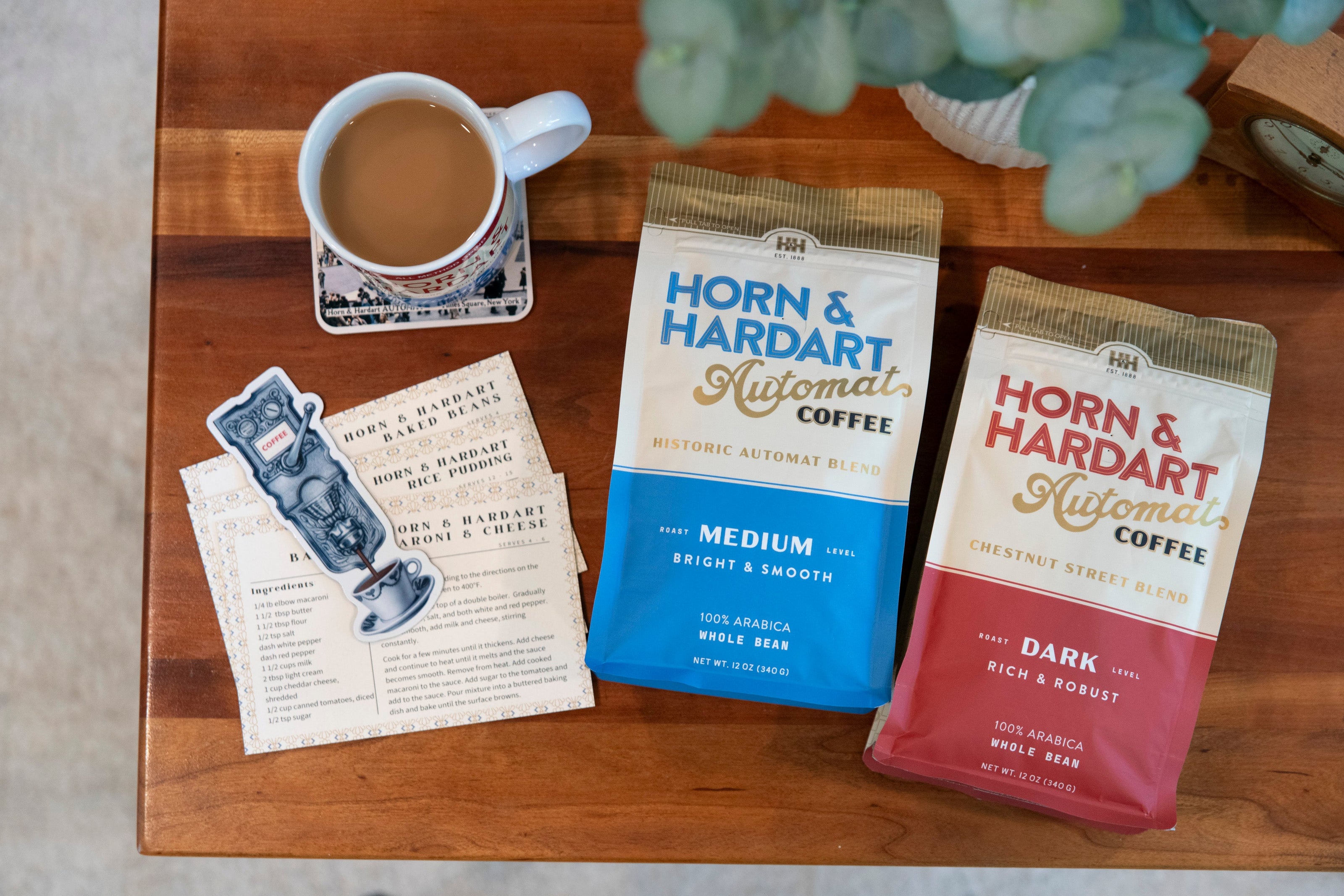 Two coffee bags from Horn & Hardart on a wooden table with a cup of coffee and a plant.