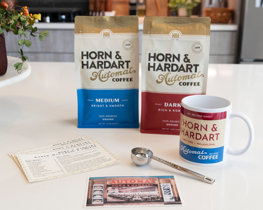 Medium roast and dark roast Automat Coffee sit on a counter with a scoop, postcard, recipe cards and all method grind mug.