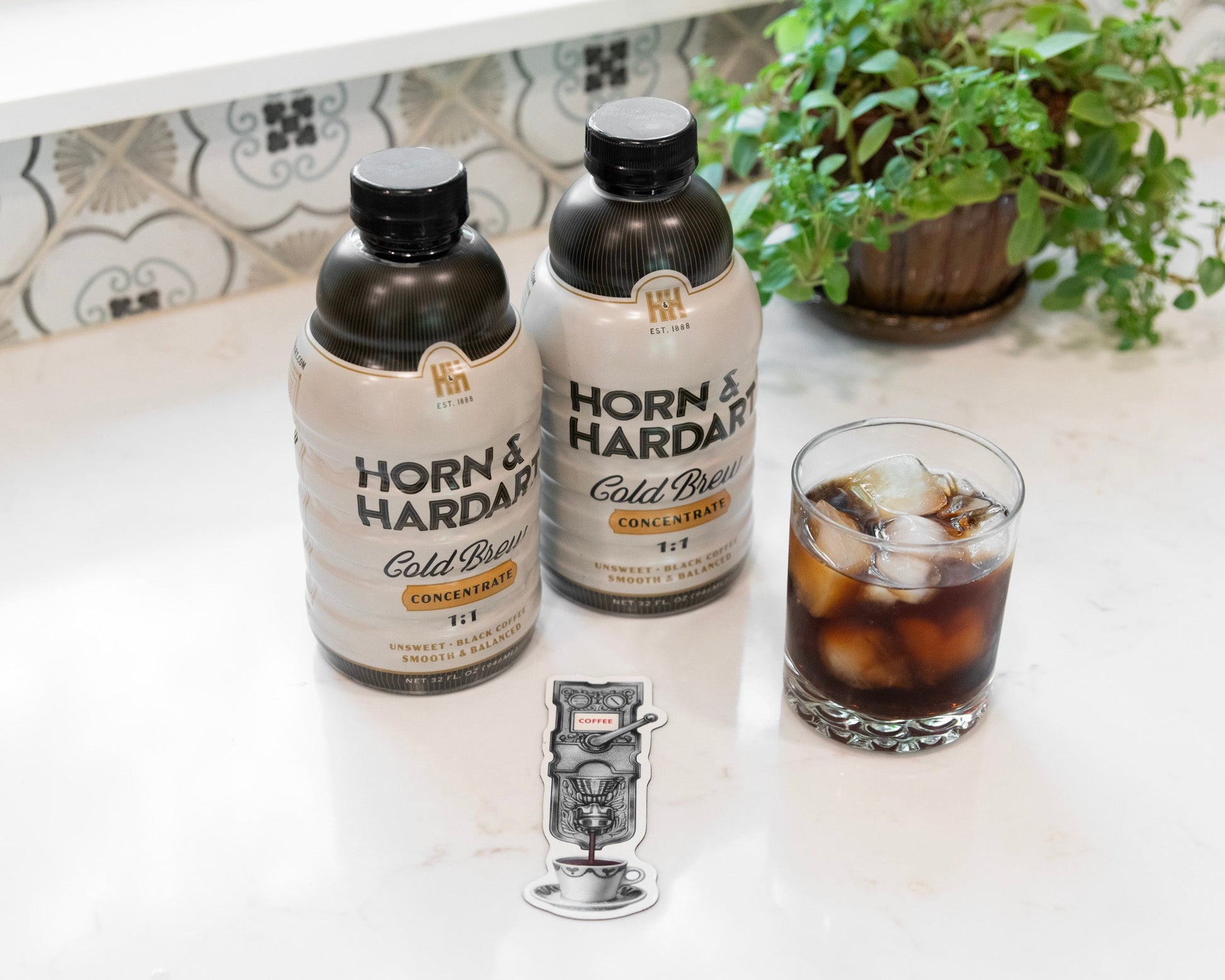 Two bottles of Horn & Hardy Cold Brew coffee with ice, a glass of iced coffee, and a small magnet on a white countertop with a tiled backsplash and a potted plant in the background.