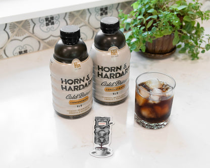 Two bottles of Horn & Hardy Cold Brew coffee with ice, a glass of iced coffee, and a small magnet on a white countertop with a tiled backsplash and a potted plant in the background.