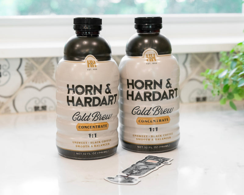 Two bottles of Horn & Hardart Cold Brew Concentrate on a white countertop in a kitchen setting, with a tiled backsplash, window light, and a potted plant in the background.