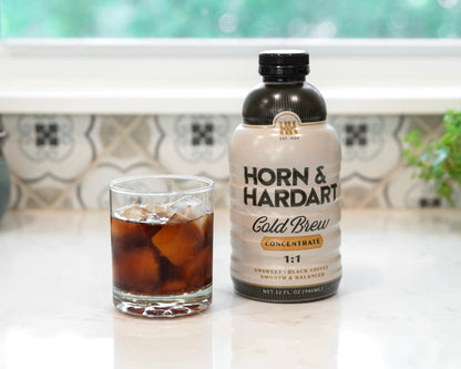 A bottle of Horn & Hardart Cold Brew coffee next to a glass of iced coffee on a white marble countertop in front of a window.