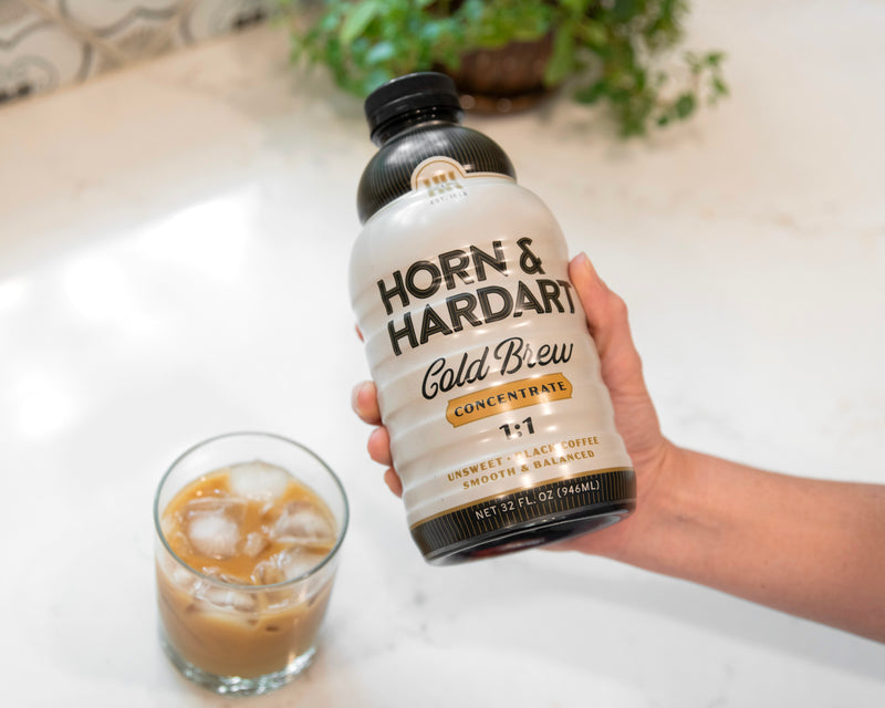 A hand holds a bottle of Horn & Hardart Cold Brew Concentrate next to a glass of iced coffee on a light marble countertop with a blurred background.