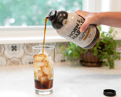 A hand pours coffee from a bottle labeled 'Horn & Hardart' into a clear glass filled with ice cubes on a kitchen counter, with a potted plant and window in the background.