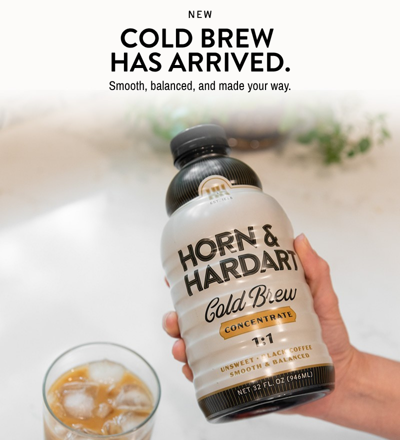 A hand holds a bottle of Horn & Hardart Cold Brew Concentrate against a bright, softly blurred background, with a glass of iced coffee and the text 'NEW COLD BREW HAS ARRIVED. Smooth, balanced, and made your way.