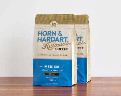 Two bags of Horn & Hardart Automat Coffee on a wooden surface against a plain white wall. The front bag is fully visible, and a second bag stands behind it slightly to the right.