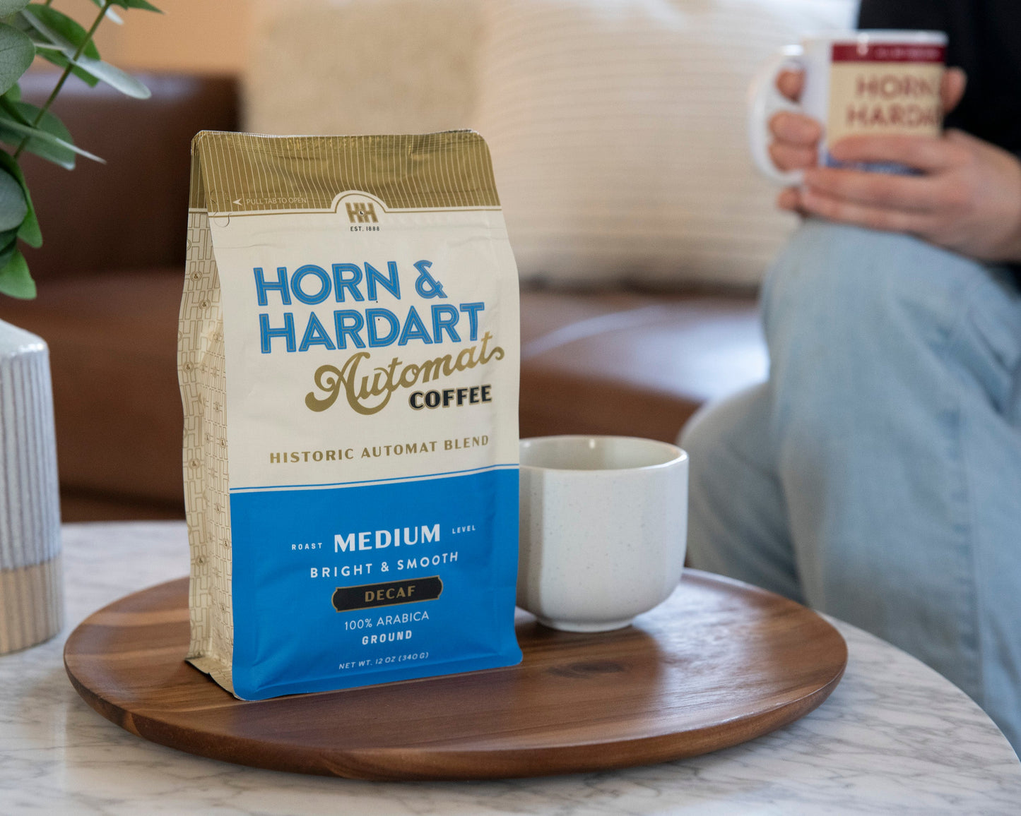 A bag of Horn & Hardart Automat Coffee sits on a round wooden tray on a table, with a white mug beside it. A person is seated in the softly blurred background holding a matching mug in a cozy indoor setting.