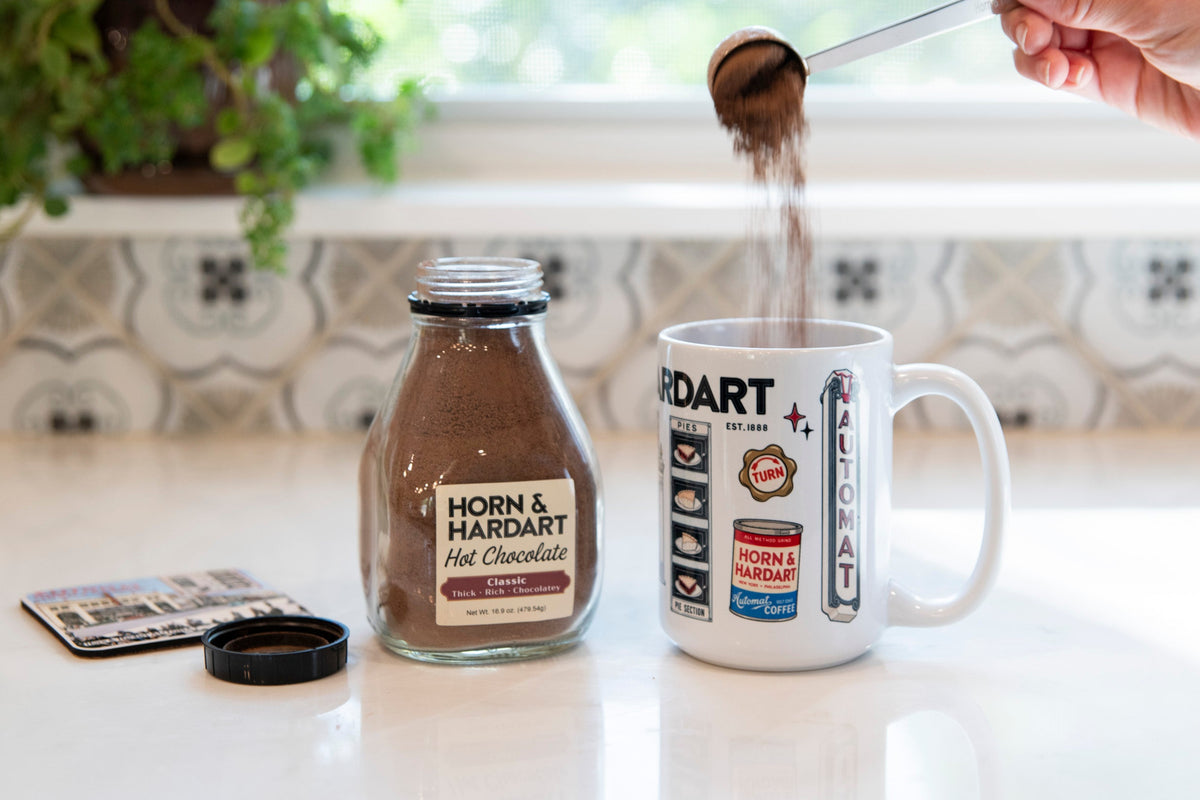 A hand pours dark brown powder from a metal scoop into a white ceramic mug printed with vintage-style railroad logos and text. A clear glass bottle labeled 'HORN & HARDART Hot Chocolate' and a small card lie on a white countertop beside the scoop in a bright kitchen setting with a window and potted plant in the background.
