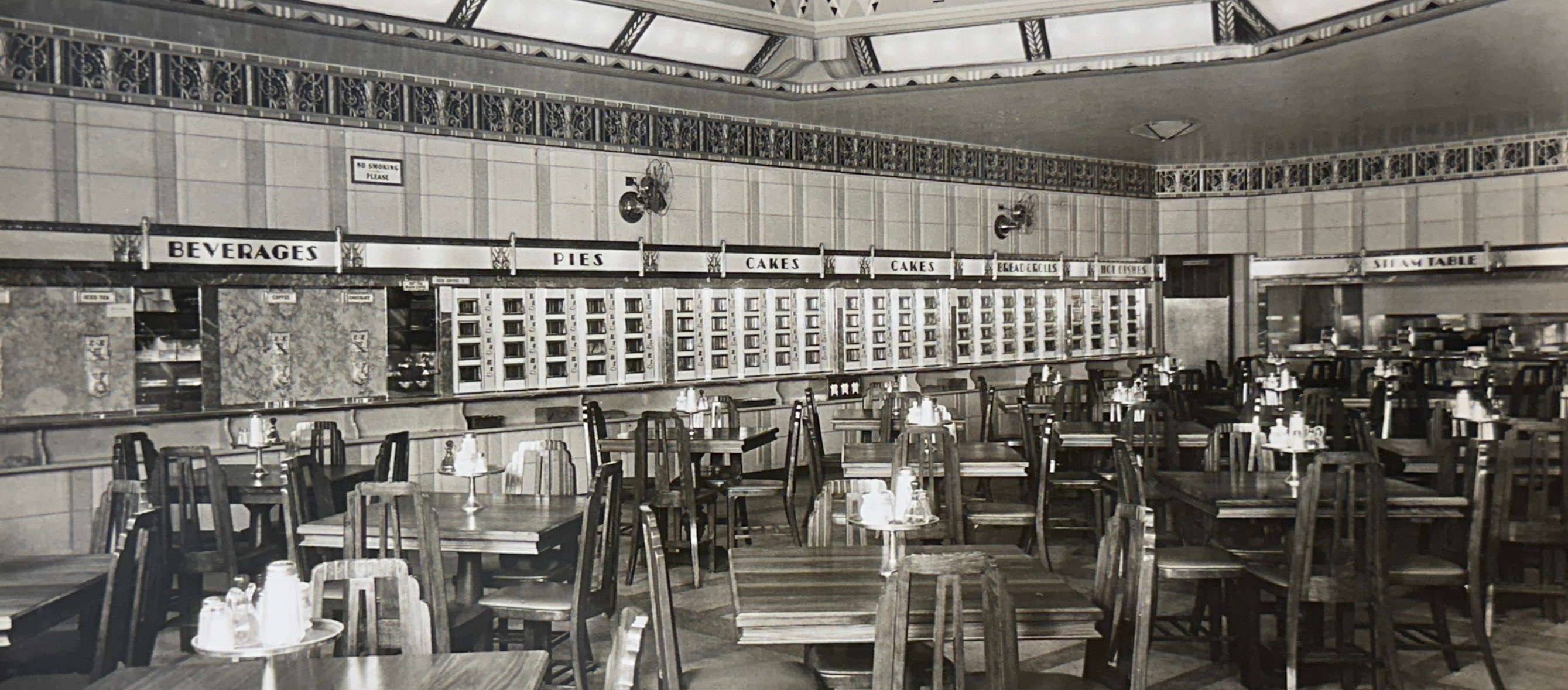 Vintage black and white photo of a diner interior with tables, chairs, and menu boards.