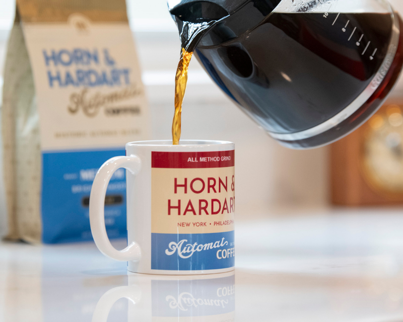 Horn & Hardart mug being filled with coffee from a carafe, with a bag of Horn & Hardart coffee in the background.