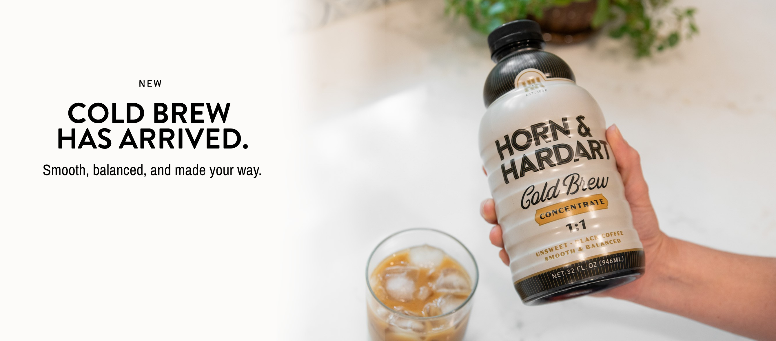 A hand holds a bottle of Horn & Hardart Cold Brew Coffee next to a glass of iced coffee on a light marble surface, with promotional text on the left.